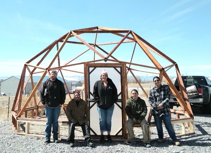 five people stand in front of a wooden geodesic dome framework