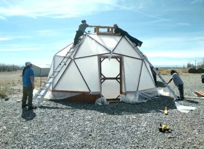 two people standing on ladders work together to install a vent on the top of a partially completed geodesic dome greenhouses. Another person looks on and a second holds the base of one ladder.