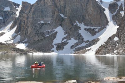 Two people boating in an alpine lake. In front of them is a cliff face with patches of snow.