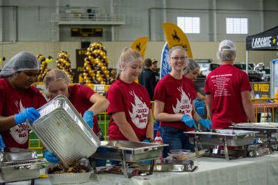 group of college students wearing hairnets and red T-shirts with 2022 Ag Day BBQ logo stand behind a buffet-style serving station in UW's indoor practice facility.