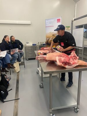 A classroom with several students watching a man in an ICMJ hat, who is holding a cut of meat and is in front of a metal table with several more cuts of meat. In the background, there is a sign that reads “Australian Pork Unlimited”. 
