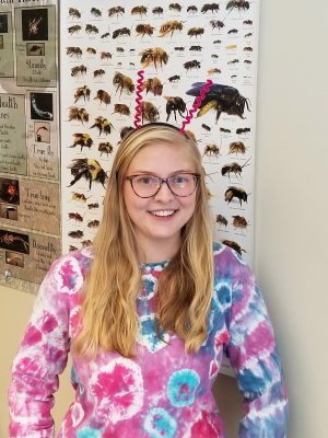 A young white woman with long blond hair and glasses wearing a headband with two "antenna". She is also wearing a pink and blue tie die shirt and is standing in front of a post with various bees on it.
