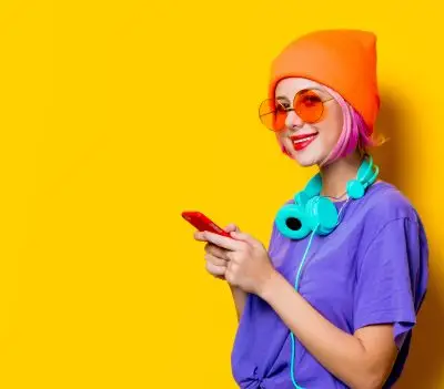 smiling teenager wearing orange beanie, round glasses with orange lenses, and a purple T-shirt. She is holding a red mobile phone and has a pair of large headphones draped around her neck.