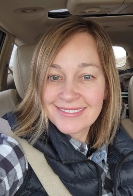 A white woman with short brown hair, blue eyes, and a puffy black vest. 