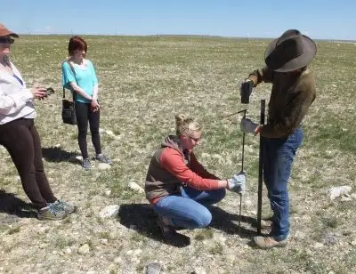 Two people watch as a person in a cowboy hat and jeans applies tools to a pole, which a woman in work clothes is holding steady.