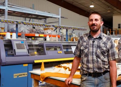 smiling man with brown beard, wearing button-up shirt and jeans, stands in front of a knitting machine and a table with a wool blanket draped over it.