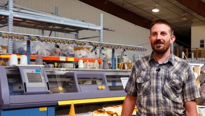 Ben Hostetler stands in front of a knitting machine and a table with a wool blanket draped over it.