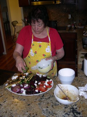 dark-haired woman wearing red shirt and yellow apron with strawberry and flower print arranges a large plate of vegetables