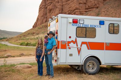 A man in a cowboy hat holding a baby and a woman stand beside a large white vehicle that reads "Shell Fire". They are parked in front of a Wyoming landscape featuring a rocky red cliff.