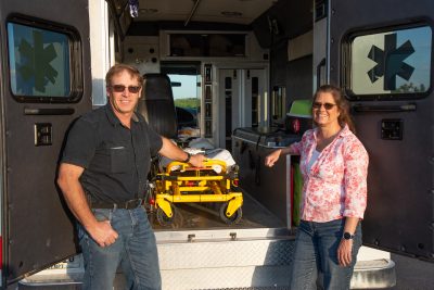 A man and a woman stand in front of the opened doors of an ambulance.