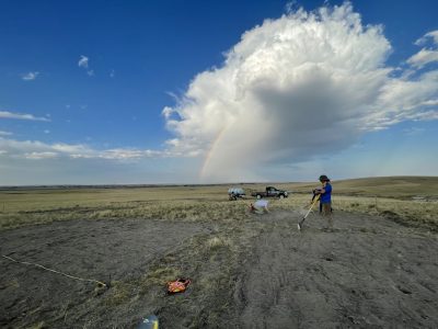 Several people working in a field. The closest person is holding a rake. The ground is dirt where the people are working; beyond them, there is more grass. The sky has a striking cloud and a rainbow. 