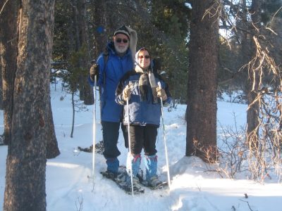 smiling man and woman wearing glasses and snow gear stand on cross country skis on a snowy trail in the woods on a sunny day