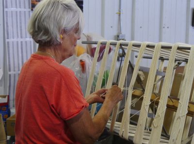 woman with short grey hair and coral-colored shirt ties strands of yarn around coils of white yarn stretched around metal bars on a machine called a skeiner. 