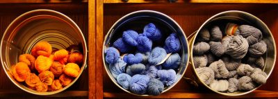 three metal buckets of yarn, one organe, one blue, and one gray, leaned sideways on a wooden shelf