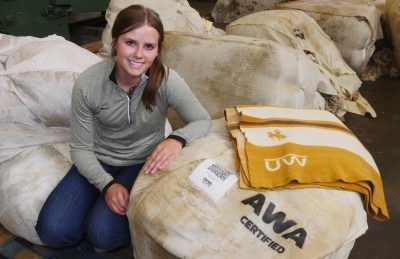 smiling woman wearing gray quarter zip and jeans kneels next to a bag of wool with a QR code and AWA certified label. A yellow, brown, and white blanket with UW logo sits on top of the bag of wool.