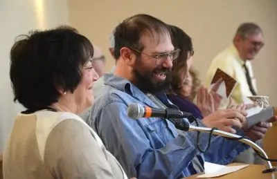 smiling woman with dark hair sits beside smiling man with dark hair and beard, also wearing glasses. A microphone is positioned on a desk in front of them.