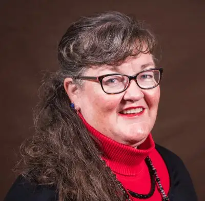 smiling woman with bangs and dark hair wearing glasses and black sweater over red turtleneck