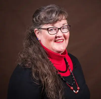 smiling woman with bangs and dark hair wearing glasses and black sweater over red turtleneck