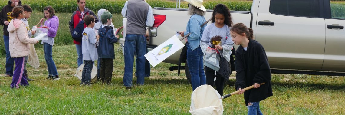 Several students and adults stand by a truck in a field, some holding several different labeled ziplock bags or nets. In the foreground, a girl is waving a large net over the grass.