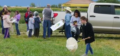 Several students and adults stand by a truck in a field, some holding several different labeled ziplock bags or nets. In the foreground, a girl is waving a large net over the grass.