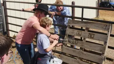 An older woman in a cowboy hat helps a kid hold a branding iron to a wood pallet while other women and kids watch.