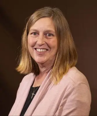 A woman with shoulder length blonde hair and a somewhat narrow face. Her skin is pale and she is wearing a pink cardigan.