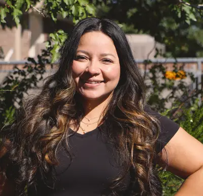 A woman with long straight black hair that curls at the ends. She has a round face, a nose ring, and somewhat olive skin. She is wearing a black t-shirt. 