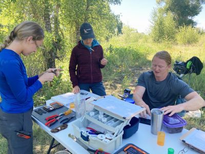 Three young adults standing around a plastic table in a forest environment. The table has several pairs of plyers, a small scale, a metal ruler and caliper, notebooks and clipboards, several containers, and a tackle box filled with several small plastic jars with lids.