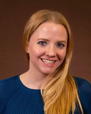 A woman with long straight blonde hair. She looks young and has pale skin. She is wearing a blue long-sleeved shirt. 