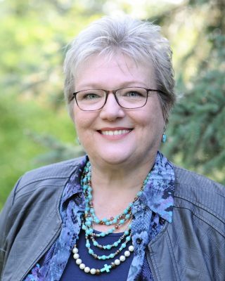 A white woman with short gray hair and glasses. She is wearing several turquoise necklaces and a blue jacket.