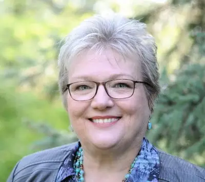 A white woman with short gray hair and glasses. She is wearing several turquoise necklaces and a blue jacket.