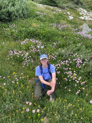 An image of a white woman wearing hiking clothes kneeling in a meadow. She has short blonde hair and large glasses.