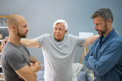 tanned old man with white hair stands between two younger men with a hand on each of their shoulders. He is smiling and the two younger men have their arms crossed but seem to have reached an agreement.