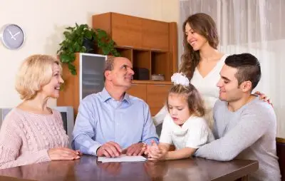 older couple, younger couple, and little girl with short hair gather around a table for a family discussion. Everyone is smiling except the little girl.