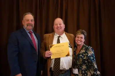 Three people with pale skin in fancy clothing. Left to right, a tall, stocky man with short gray hair and a handlebar mustache. A stocky, balding man holding up an award. He is wearing a brown suit jacket and tie, a white shirt with UW on the lapel, and jeans with a large belt buckle. A shorter woman with styled short brown hair. 