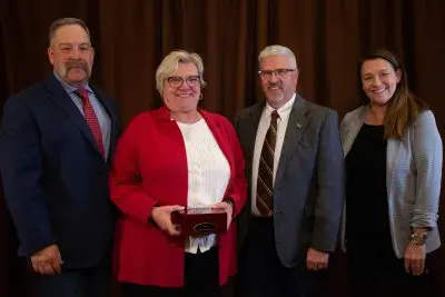 Four people in fancy clothing. All have pale skin. From left to right, a tall, stocky man with short gray hair and a handlebar mustache. A woman with short gray hair and glasses holding a small box; she is wearing red. A man with short white hair, a mustache and beard, and glasses. A woman with long brown hair.