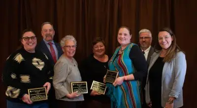 Seven adults in fancy clothing. From left to right, a stockier woman with long brown hair, pale skin, and glasses, holding an award. A tall, stocky man with gray hair and a handlebar mustache and pale skin. A short woman with short gray hair, glasses, and pale skin, holding an award. A shorter fat woman with short brown hair and pale skin, holding an award. A tall stocky woman with long brown hair wearing a bright blue dress holding an award. A man with white hair, a mustache and beard, pale skin, and glasses. And a woman with long brown hair and pale skin.