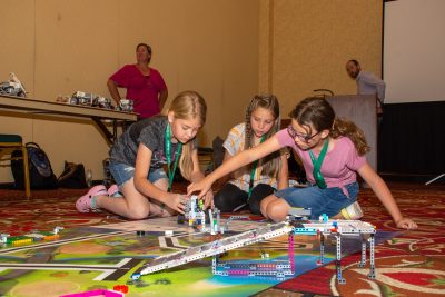Three girls sitting on the floor working on some Lego robots. The robot is on a mat and in front of them is a lego ramp. The girl on the right is reaching over to the robot, while the girl on the left holds it and the girl in the middle looks on.