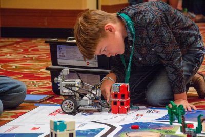 A boy leaning down over a lego robot. The robot has pincers and wheels and is right next to a small red lego tower. The boy is white with short brown hair and is wearing a green lanyard.