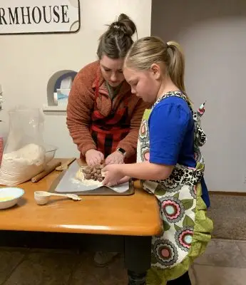 woman and young girl wearing aprons prepare food on a baking sheet placed on a wooden table