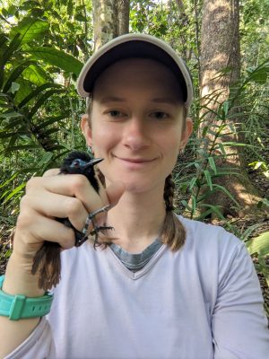 A small black bird held in the hand of a white woman. Its feet, head, and tail stick out from her grip. She is wearing a baseball cap, a white athletic long sleeve, and a bright teal watch, has shoulder length brown braids, and is smiling. The background has tropical plants and trees.
