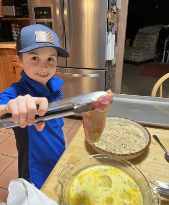 smiling boy wearing blue baseball cap holds a piece of raw meat in tongs, ready to dip in a dry seasoning mix after being coated with raw egg