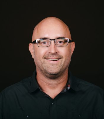 An image of a bald white man with rectangular glasses and some stubble. He is wearing a dark button up shirt.