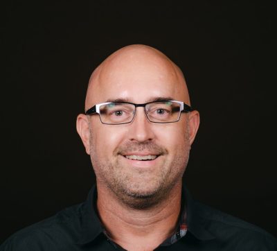 An image of a bald white man with rectangular glasses and some stubble. He is wearing a dark button up shirt.