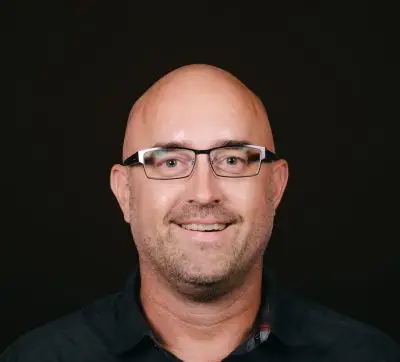 An image of a bald white man with rectangular glasses and some stubble. He is wearing a dark button up shirt.