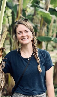 A young, thin, white woman with a long blonde braid. She has on a nice t-shirt, sunglasses on her head, brown wooden earrings, and a necklace. The background is filled with plants which look like banana trees.