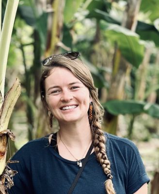 A young, thin, white woman with a long blonde braid. She has on a nice t-shirt, sunglasses on her head, brown wooden earrings, and a necklace. The background is filled with plants which look like banana trees.