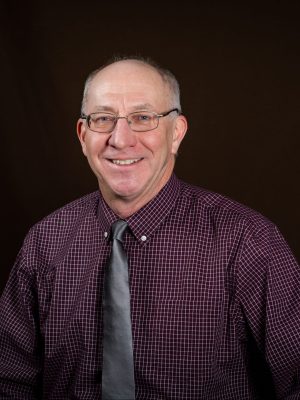 An older white man wearing wire rim glasses and a checkered wine-colored button up shirt and a gray tie. He has short gray hair and is clean-shaven and balding. 