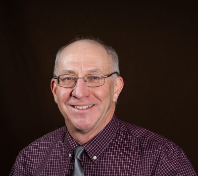 An older white man wearing wire rim glasses and a checkered wine-colored button up shirt and a gray tie. He has short gray hair and is clean-shaven and balding.