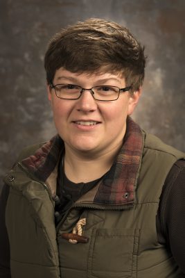A white woman with short straight brown hair wearing a warm vest with flannel collar, brown shirt, and square glasses. 
